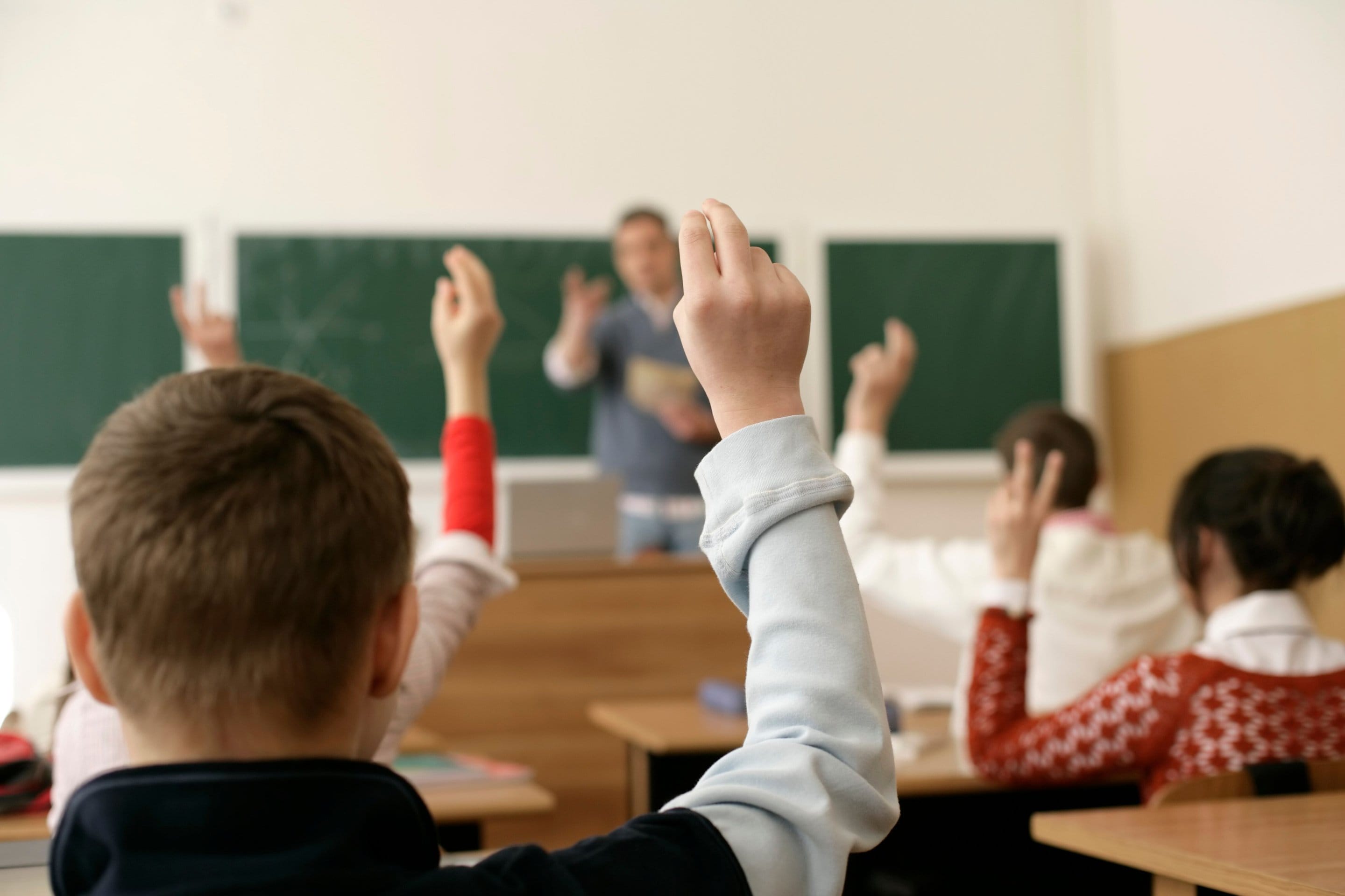 Elevi cu mâinile ridicate într-o clasă, văzuți din spate, cu profesorul la tablă